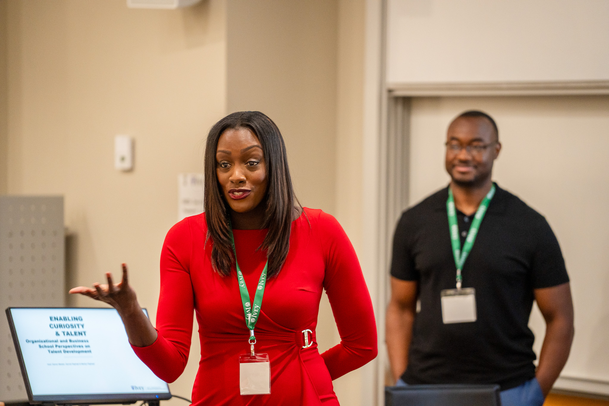 ICON Talent partners cofounders Marilyn & Derrick Raphael presenting at the Wood Symposium 2024. Marylin is in the front of the photo, wearing a red dress, leading an interactive session. Derrick is in the background, slightly out of focus, he is wearing jeans and a black teeshirt, has his hands in his pockets and is smiling in agreement with Marilyn's comments.