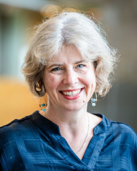 Headshot of Maggie Weller, smiling. She is wearing a blue top, a silver necklace, and dangling blue bead earrings. Her silver hair is styled in an ear-length bob.
