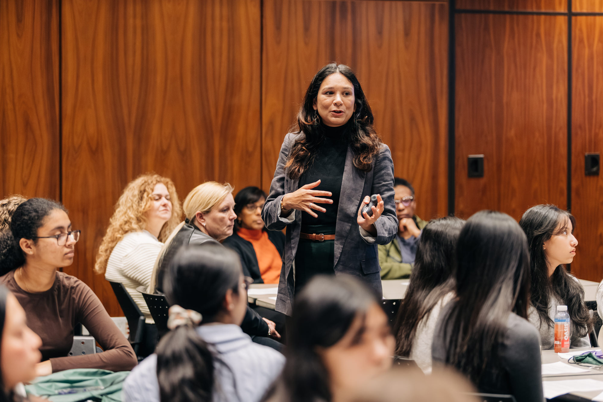 Barnini Bhattacharyya stands in the middle of a room filled with GEMinar participants. Wood paneling lines the wall behind her. Participants, seated at tables of eight, are actively engaged in discussion