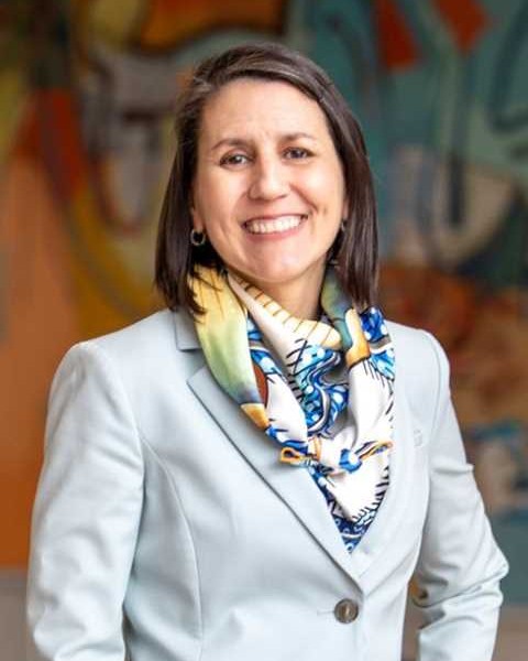 Zoe Kinias, smiling, stands in front of a large, colorful artwork in the Ivey Building. She is wearing a light blue jacket and a blue-and-green scarf.