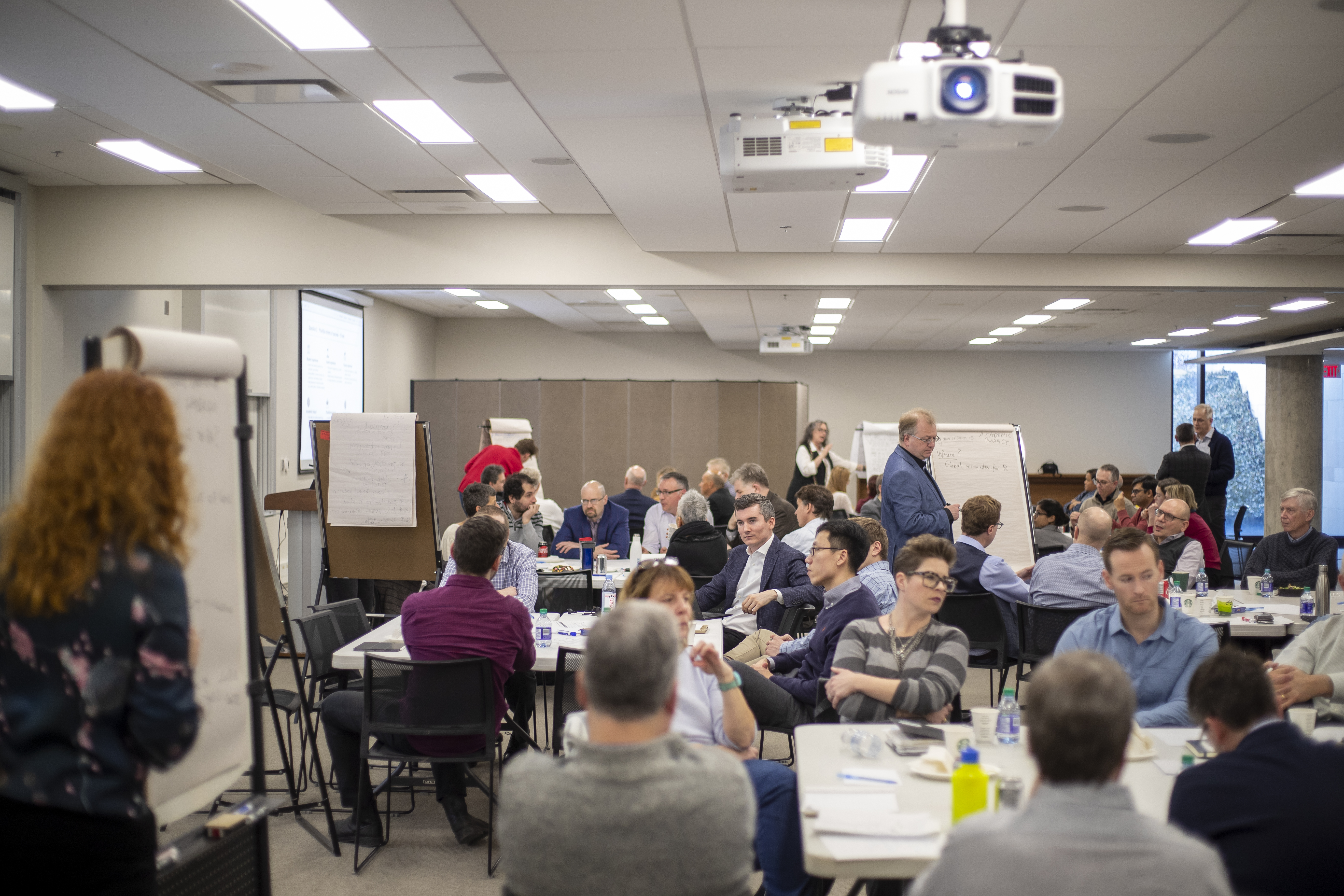 A room filled with Ivey professors, sat around tables with a flip chart at each table.  The professors are all engaged in discussions
